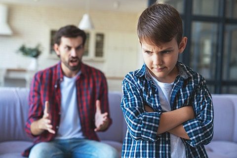 An angry father quarrels with his little son, who stands offended in the foreground at his home in the living room.