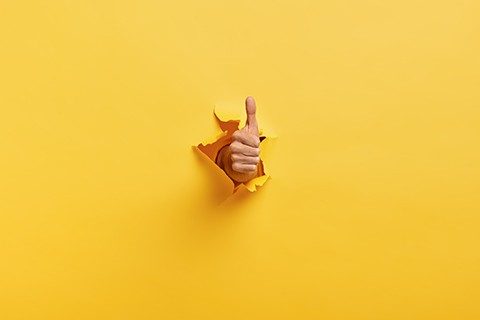 Image of unrecognizable man makes thumb up gesture, demonstrates approval or agreement, gestures through torn paper wall yellow background. Body language concept. Hand sign. Hole in wall. Like gesture