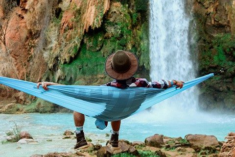A wide shot of a male lying on a hammock beside a waterfall flowing down from a hill