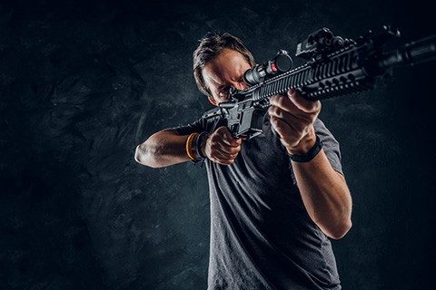 Crazy middle-aged man dressed in casual clothes holding an assault rifle and aims at the target. Studio photo against a dark textured wall