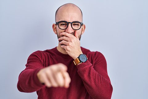 Young bald man with beard standing over white background wearing glasses laughing at you, pointing finger to the camera with hand over mouth, shame expression